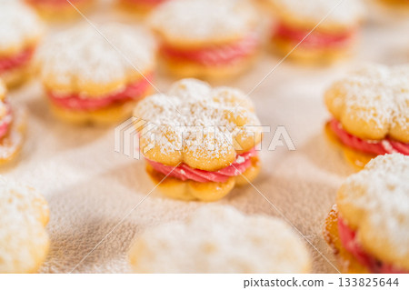 Close-up side view of a lemon spritz cookie sandwich filled with cranberry buttercream. The golden cookie is dusted with powdered sugar, and the vibrant pink buttercream adds contrast. The background 133825644