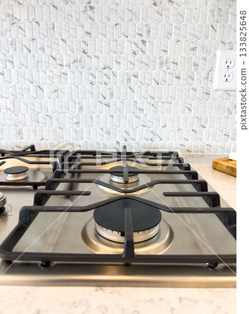 A gas cooktop is positioned in front of a white stone tile backsplash with a power outlet nearby. The image captures a clean and modern culinary space. 133825648