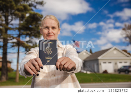 Woman standing outdoors and holding a United States passport toward the camera, with houses, trees and a blue sky in the background. 133825782