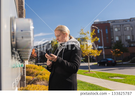 Middle-aged woman checking electricity meters outside a residential building and entering usage data on her smartphone during a sunny autumn day Middle-aged woman checking electricity meters outside a residential building and entering usage data on her smartphone during a sunny autumn day 133825787