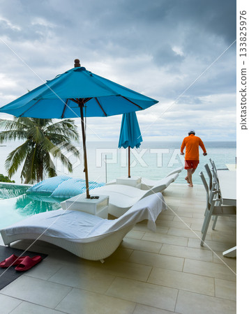 it is raining heavily in the background, a man near the pool is relaxing and watching the rain in the sea, a palm tree by the pool in a luxury villa and the sea it is raining heavily in the background, a man near the pool is relaxing and watching the rain in the sea, a palm tree by the pool in a luxury villa and the sea 133825976