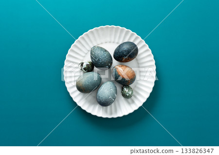 Decorative Easter eggs with various textures and colors are arranged neatly on a white plate, resting on a vibrant teal surface, creating a festive spring atmosphere Decorative Easter eggs with various textures and colors are arranged neatly on a white plate, resting on a vibrant teal surface, creating a festive spring atmosphere 133826347