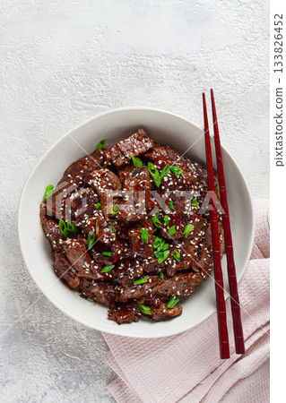 Crispy beef, with green onions and sesame seeds, in a bowl with chopsticks, on a textured surface, on a light background, no people Crispy beef, with green onions and sesame seeds, in a bowl with chopsticks, on a textured surface, on a light background, no people 133826452