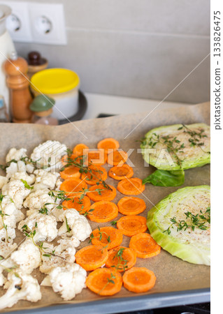 Cauliflower, carrots, and cabbage are sliced and arranged on a tray lined with parchment paper, ready for roasting with herbs in a warm kitchen setting 133826475