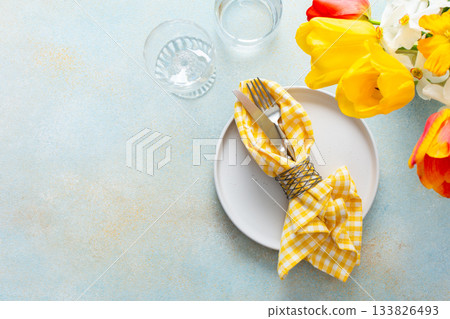 A charming table setting for spring includes a white plate with a yellow checkered napkin, fork, and knife neatly arranged among colorful flowers in bloom 133826493
