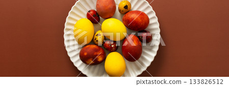 A collection of brightly colored Easter eggs in various sizes and patterns is displayed on a white scalloped plate, resting on a warm brown surface, banner 133826512