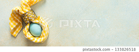 A pale blue Easter egg is beautifully wrapped in a yellow checkered cloth, tied with a silver ring on a soft, light-colored surface. This represents festive spring decorations, banner 133826518