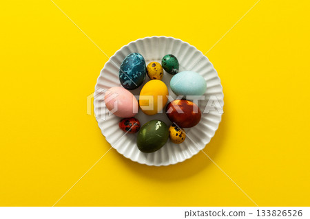 A variety of beautifully painted Easter eggs in different colors and patterns are placed on a delicate white plate. The vibrant hues stand out against the cheerful yellow backdrop A variety of beautifully painted Easter eggs in different colors and patterns are placed on a delicate white plate. The vibrant hues stand out against the cheerful yellow backdrop 133826526