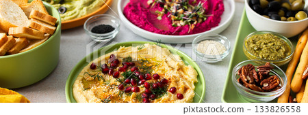 A vibrant spread showcases different types of hummus including classic, beetroot, broccoli, and turmeric. Accompanied by olives and breadsticks, it invites sharing and enjoyment, banner A vibrant spread showcases different types of hummus including classic, beetroot, broccoli, and turmeric. Accompanied by olives and breadsticks, it invites sharing and enjoyment, banner 133826558