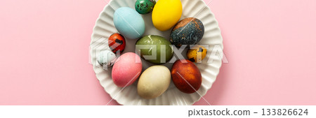 A collection of vibrant Easter eggs in various colors is artfully arranged on a white plate, set against a soft pink background, capturing the essence of the holiday's decorations, banner 133826624