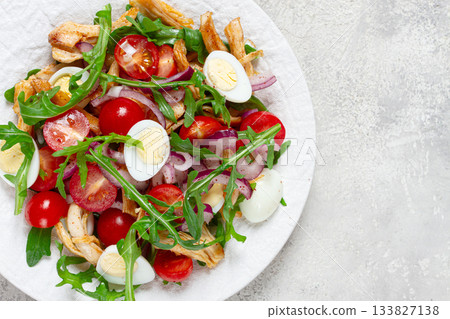 a warm salad with fried chicken fillet, arugula and cherry tomatoes, and boiled quail eggs, on a white plate, no people. 133827138