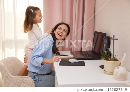 Happy mother working from home laughing while child plays and distracts her Happy mother working from home laughing while child plays and distracts her 133827849