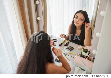 Woman applying mascara to her eyelashes in front of a makeup mirror Woman applying mascara to her eyelashes in front of a makeup mirror 133827861