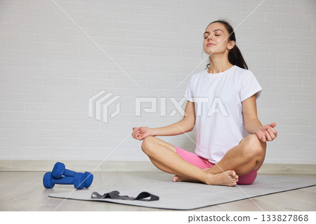 Woman sitting in lotus pose on a mat, meditating after exercise session at home 133827868