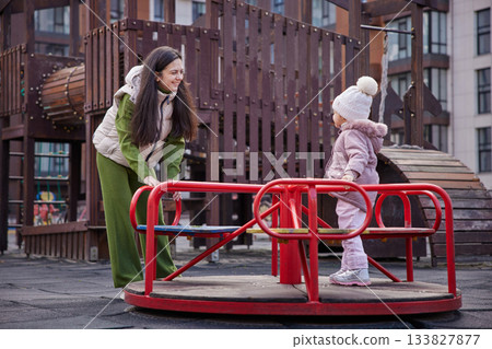 Mother and daughter playing together on a merry-go-round in an urban park Mother and daughter playing together on a merry-go-round in an urban park 133827877