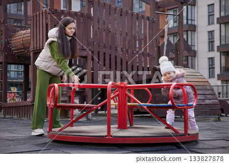 Mother and daughter playing together on a merry-go-round in an urban park Mother and daughter playing together on a merry-go-round in an urban park 133827878