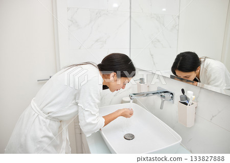 Woman in bathrobe rinsing face with water from a modern sink Woman in bathrobe rinsing face with water from a modern sink 133827888