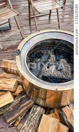 Modern stainless steel smokeless fire pit surrounded by firewood on a wet wooden deck with outdoor chairs 133828588
