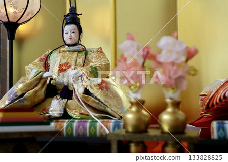 An elegant photo of a male Hina doll, the Emperor and Empress, standing out against a gold screen with peach blossoms in the foreground. Material for a Japanese-style spring event. 133828825