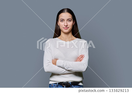 Beautiful young woman with crossed arms looking at camera on grey background 133829213