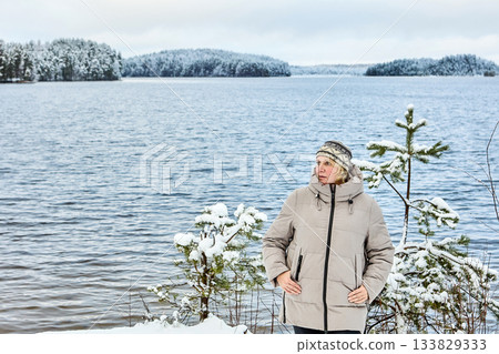 Woman stands by tranquil lake, gazing into distance, clad in winter jacket, amid snow-covered evergreens. Woman stands by tranquil lake, gazing into distance, clad in winter jacket, amid snow-covered evergreens. 133829333