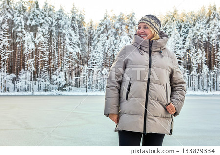 Smiling stands mature white woman in warm winter coat and patterned knit hat on snowy shore during winter day near frozen lake in northern forest. Smiling stands mature white woman in warm winter coat and patterned knit hat on snowy shore during winter day near frozen lake in northern forest. 133829334