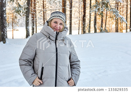 Warmly dressed mature woman stands in snowy forest while her companion takes photo. Warmly dressed mature woman stands in snowy forest while her companion takes photo. 133829335