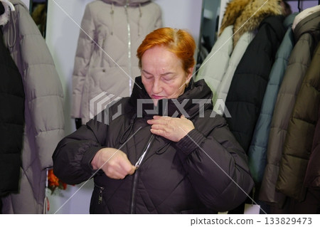 Young woman in new black leather jacket looking at herself in mirror in fitting room in boutique while standing next to her husband Young woman in new black leather jacket looking at herself in mirror in fitting room in boutique while standing next to her husband 133829473
