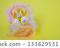 On a yellow background, Easter eggs with bunny ears in a basket of feathers. Easter concept. Space for your text. 133829531