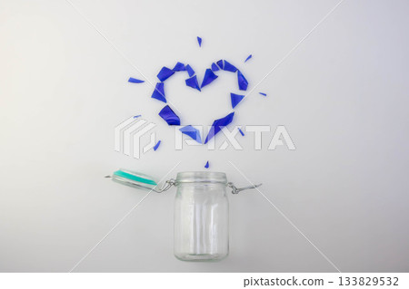 A small glass jar with a lid and a beautiful heart made of blue glass shards on a white background. The concept of love and separation. A small glass jar with a lid and a beautiful heart made of blue glass shards on a white background. The concept of love and separation. 133829532
