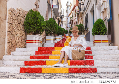 Young woman in a white dress sitting on the colourful Spanish flag stairs in the old town of Calpe, Spain 133829778