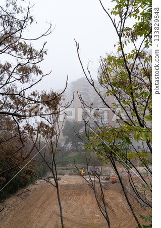 Construction vehicles sit on a dirt lot surrounded by trees on a foggy day with buildings in the background captured in Zaragoza, Spain. Construction vehicles sit on a dirt lot surrounded by trees on a foggy day with buildings in the background captured in Zaragoza, Spain. 133829848