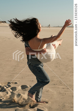 Man carrying woman in his arms on sandy beach, her hair flowing in the wind during a romantic and playful moment. Man carrying woman in his arms on sandy beach, her hair flowing in the wind during a romantic and playful moment. 133830326