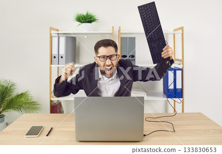 Angry shouting man working on workplace in office trying to destroy the monitor with his keyboard. Angry shouting man working on workplace in office trying to destroy the monitor with his keyboard. 133830653