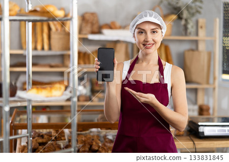 Portrait of female baker with mobile phone in hands in bakery interior Portrait of female baker with mobile phone in hands in bakery interior 133831845