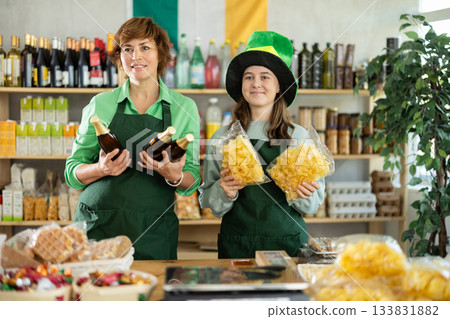 Two female sellers posing in green costume and hat, offering bottle of irish ale in store decorated for St. Patrick Day Two female sellers posing in green costume and hat, offering bottle of irish ale in store decorated for St. Patrick Day 133831882