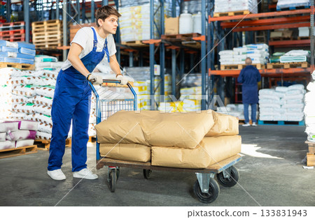 In warehouse of store, stevedore loader young man pushes and carries large cart for bulky cargo In warehouse of store, stevedore loader young man pushes and carries large cart for bulky cargo 133831943