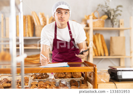 Young baker reading documents and writing notes on paper in the interior of private bakery Young baker reading documents and writing notes on paper in the interior of private bakery 133831953