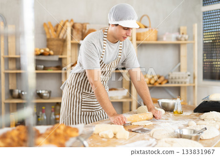 Bakery worker, young baker, rolls out raw yeast dough with rolling pin 133831967