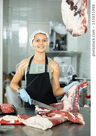 Positive young saleswoman cutting piece of meat in butcher shop 133831998