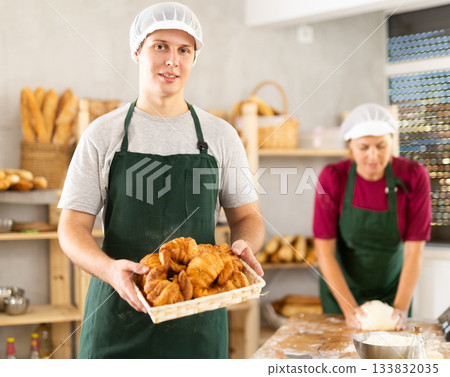 Young baker in uniform stands in small private bakery offering hot croissants 133832035