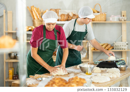 Two professional bakers in apron standing at work table and kneading dough, cutting into pieces and weighing for baking bread in bakery Two professional bakers in apron standing at work table and kneading dough, cutting into pieces and weighing for baking bread in bakery 133832129