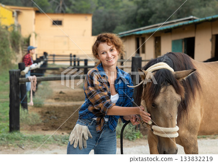 Happy woman farmer in plaid shirt and gloves preparing horse for riding and walking in pasture on farm Happy woman farmer in plaid shirt and gloves preparing horse for riding and walking in pasture on farm 133832219
