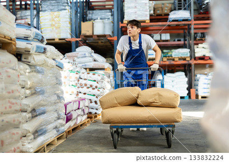 In warehouse of store, stevedore loader young man pushes and carries large cart for bulky cargo 133832224