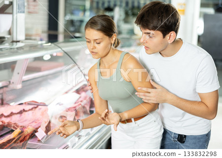 Young man and woman purchasers buying meat in butcher shop Young man and woman purchasers buying meat in butcher shop 133832298