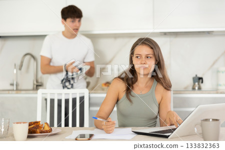 Young girl sitting at the table while her boyfriend is quarreling with her standing behind Young girl sitting at the table while her boyfriend is quarreling with her standing behind 133832363