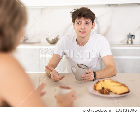 Adult woman and young guy drink tea and talk in kitchen Adult woman and young guy drink tea and talk in kitchen 133832390