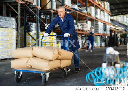 In warehouse rack area of store, loader mature man pushes and carries large cart for bulky cargo In warehouse rack area of store, loader mature man pushes and carries large cart for bulky cargo 133832442