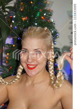 A young blonde model looks at the camera, smiles and rejoices. In the background is a New Year's tree and garland. Portrait photography. 133833095