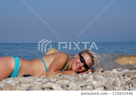 A blonde in glasses and a swimsuit is looking at the sun, sunbathing, and laughing. She is lying on the shore of Lake Baikal. A blonde in glasses and a swimsuit is looking at the sun, sunbathing, and laughing. She is lying on the shore of Lake Baikal. 133833446